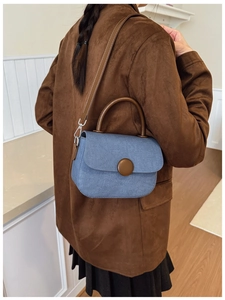 Detail of blue denim crossbody bag with brown leather strap worn by a model in a cozy room