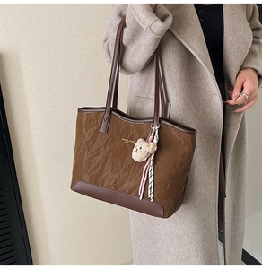 Detail of brown pleated tote bag featuring a plush bear charm and leather strap accents