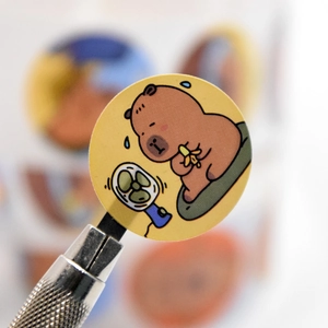Vista macro di un simpatico adesivo capibara tenuto con una pinzetta metallica di precisione