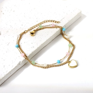 Double-layered gold anklet with colorful beads and a heart pendant on a stone surface