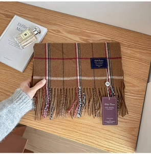 Detail shot of the brown plaid wool scarf showing soft fringe and label on a desk