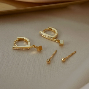 Detail of gold cube charm earrings and ball studs set on warm textured fabric