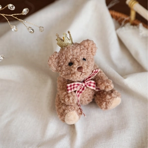 Brown curly plush teddy bear with gold crown and red gingham bow on white background