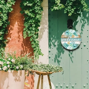 Round wooden welcome door sign with teal floral design hanging on a green cottage door