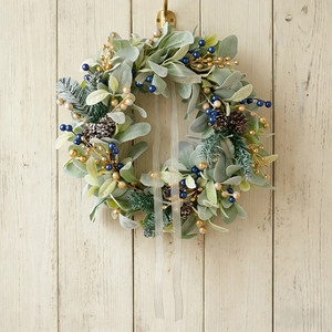 Christmas wreath with flocked leaves, blue berries, and gold ribbons on white wooden wall
