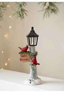 Close-up of Christmas resin ornament featuring a lit street lamp, birds, and Merry Christmas sign