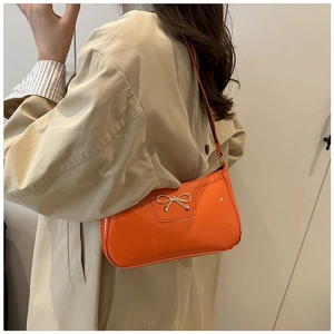 Detail of orange patent leather shoulder bag featuring a gold bow buckle on a model