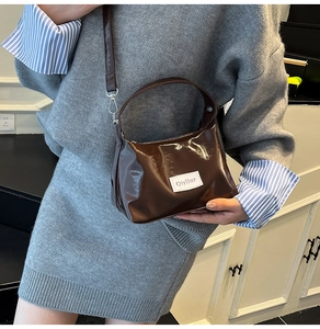 Dark brown glossy shoulder bag with white logo patch shown on a model for casual styling