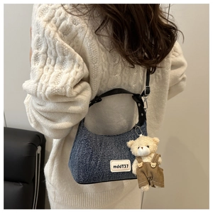 Detail of blue denim shoulder bag featuring a white logo patch and a cute bear bag charm