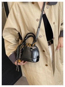 Detail of black glossy shell bag with gold hardware and woven bell charm on a trench coat