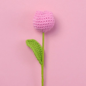 Close-up of a handmade pink crochet tulip flower with green stem on a soft pink background