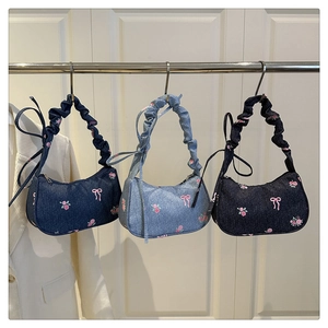 Three denim shoulder bags with floral embroidery and ruched handles hanging on a rack