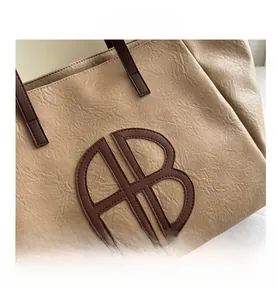 Detail einer beigen Tragetasche mit Faltenstruktur und braunem Leder-AB-Logo