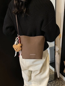 Matte braune Beuteltasche mit Bärenanhänger und weißem Slogan an einem Crossbody-Modell