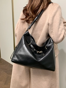 Large black polished leather tote bag shown on model wearing a beige coat in minimalist setting