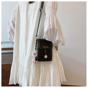Detail of a black mini crossbody bag with gold buckle and a pink plush charm on a white dress