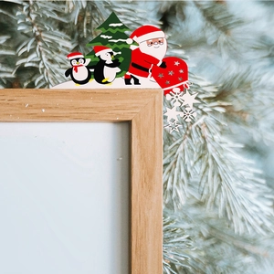 Detail of wooden Santa and penguin frame decor with snowflakes and pine background