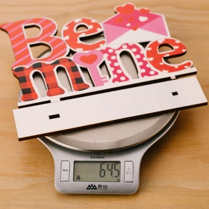Détail d'un panneau en bois 'Be Mine' pesant 645g sur une balance numérique