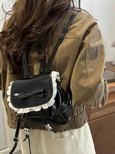 Black patent leather mini backpack with white lace trim and bow shown on a model