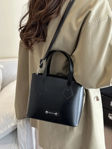 Classic black trapezoid handbag with silver metal tag shown on a female model with tan coat
