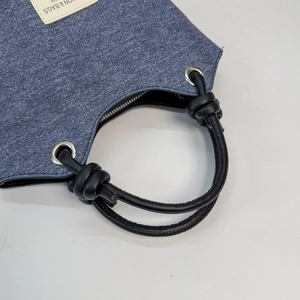 Dettaglio borsa a secchiello in denim blu con manico in pelle nera annodato e occhielli