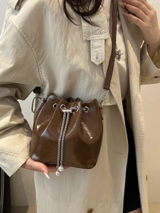 Minimalist deep brown patent leather bucket bag with pearl chain detail worn by a model