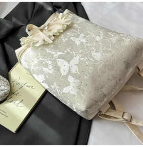 Detail of cream backpack with silver butterfly patterns, ruffles, and a pearl bow