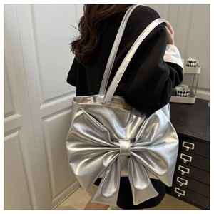 Silver metallic tote bag with large bow detail shown on shoulder in minimalist interior