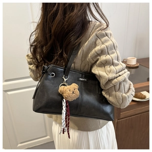 Detail of black tote bag with brown plush bear charm and woven tassel shown on shoulder