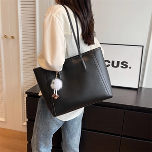 Side view of a large black leather tote bag with a white plush doll charm on a model