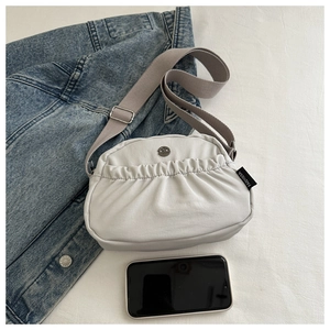 Close-up of pleated canvas crossbody bag with metal logo and wide strap next to a mobile phone