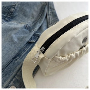 Detail of cream pleated crossbody bag featuring a round metal logo and black zipper