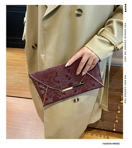 Detail of burgundy snakeskin clutch with gold metal trim held by a model in a trench coat