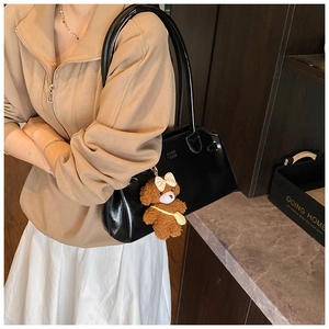 Glossy black leather tote bag with a brown plush bear charm worn with casual beige outfit