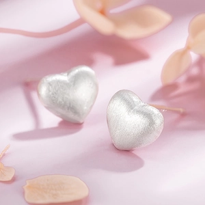 Macro detail of silver heart stud earrings with a brushed matte finish on a pink background