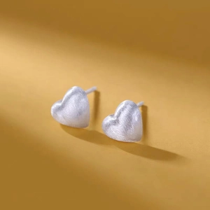 Brushed silver heart stud earrings shown at a 45-degree angle on a mustard yellow background