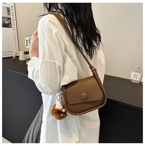 Brown semi-circular leather shoulder bag with plush pendant shown on a white shirt model