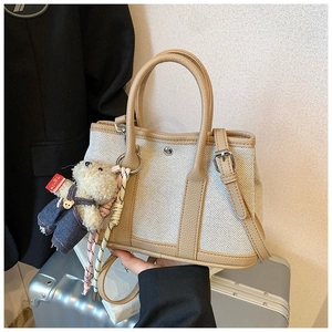 Canvas bucket tote bag with leather trim and teddy bear charm held in front of suitcase