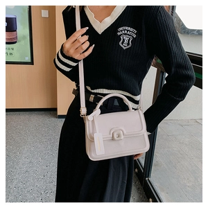 Model wearing a small beige square crossbody bag with handle in a cozy cafe setting