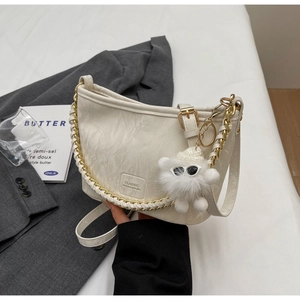 Detail van gebroken witte crossbodytas met goudkleurige ketting en pluche bedeltje op bed