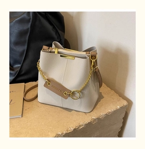 Detail of cream pebbled leather bucket bag with gold chain and brand tag on stone surface