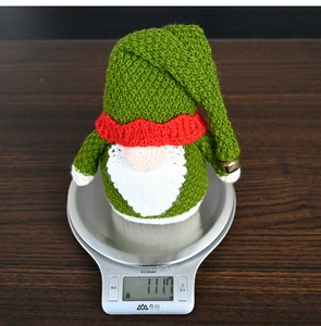 Macro shot of a 111.7g green knitted Christmas gnome on a scale showing fabric texture