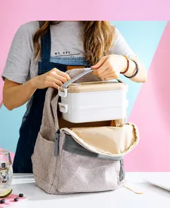 White double-layer bento box with wood lid being placed into a beige backpack