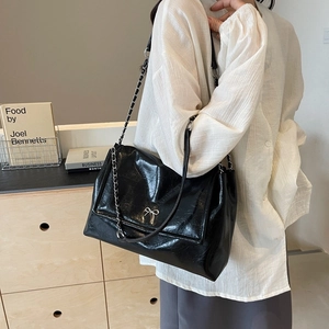Black glossy tote bag with silver bow detail and chain strap shown on model shoulder