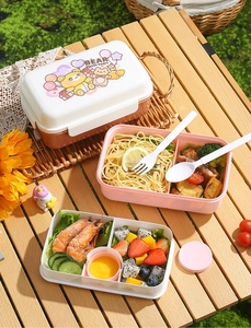Outdoor picnic scene showing a pink and white double-layer lunch box with cutlery