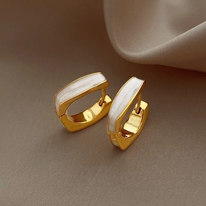 Detailed view of gold rectangular huggie earrings with white enamel inlay on beige stand
