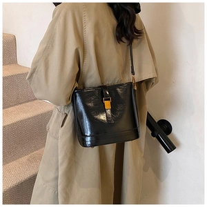 Détail d'un sac seau en cuir verni noir avec boucle dorée sur un trench-coat kaki