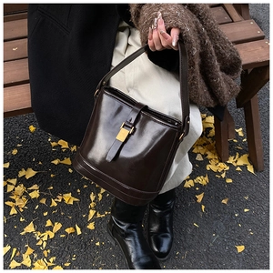 Détail d'un sac seau en cuir ciré marron foncé tenu par un mannequin sur un banc de parc