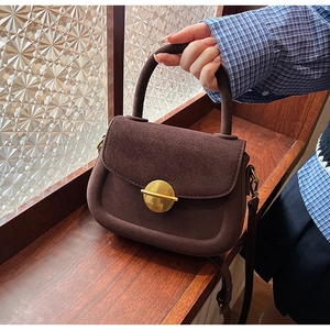 Detail of coffee brown suede saddle bag with a round gold metal buckle on wooden windowsill