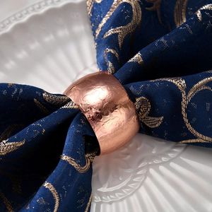Portatovagliolo oro rosa con texture martellata su tessuto blu scuro ricamato
